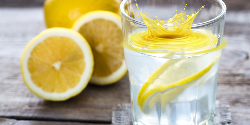 A glass of lemon water on a table with lemons next to it and a graphic making it look like a lemon splash coming out of the glass.