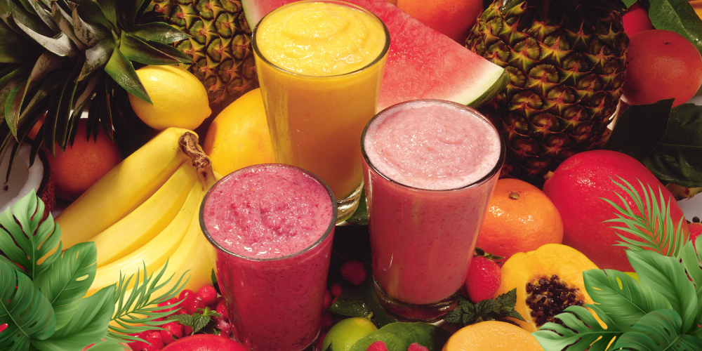 3 smoothies around a bunch of tropical fruit with grass in each corner.